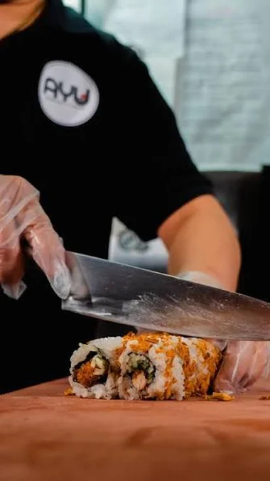 Mitarbeiter im Ayu-Sushi-Shirt schneidet mit einem langen Messer eine Inside-out-Rolle